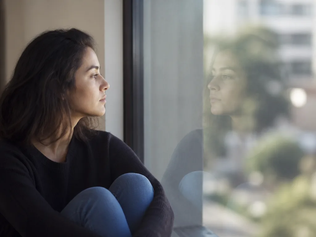 Person sitter vid ett fönster och tittar eftertänksamt på sin egen spegling i glaset